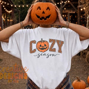 May include: A white short-sleeved t-shirt featuring a graphic of an orange jack-o'-lantern pumpkin centered between the letters C, O, Z, and Y, which are filled with an orange and black checkerboard pattern, with the word season below.