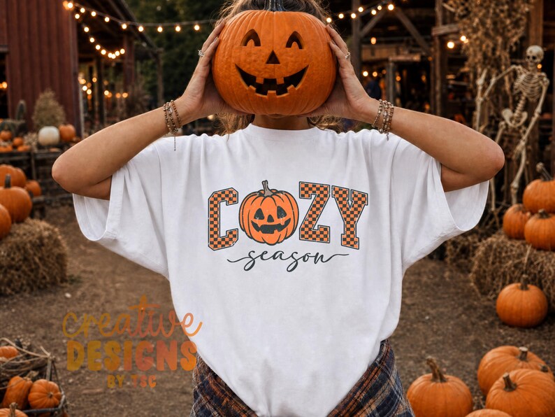 May include: A white short-sleeved t-shirt featuring a graphic of an orange jack-o'-lantern pumpkin centered between the letters C, O, Z, and Y, which are filled with an orange and black checkerboard pattern, with the word season below.