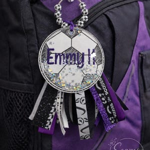 May include: A soccer-themed backpack charm with a white and black soccer ball design. The charm features the name "Emmy K" in purple glitter and is adorned with ribbons in black, white, and purple. The charm is attached to a black and purple backpack.