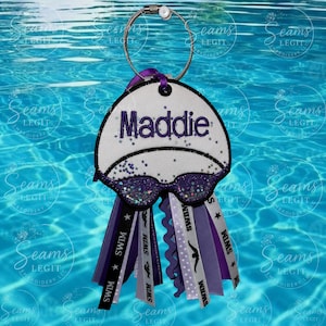 May include: A personalized swim team keychain featuring a white swim cap design with the name "Maddie" in purple. The keychain includes purple glitter goggles and ribbons with "SWIM" text and swimming figures. The background is a blue pool.