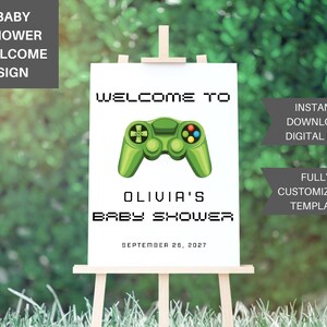 May include: A white rectangular welcome sign on a wooden easel featuring a green video game controller graphic. The text reads: WELCOME TO OLIVIA'S BABY SHOWER SEPTEMBER 26, 2027. The design uses a pixelated font style for the main headings and a simple sans-serif font for the date.