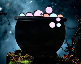 May include: A black cauldron with a rim and handles, filled with pink, blue, and orange bubble-like shapes. The cauldron is sitting on a mossy surface in front of a dark, foggy background.