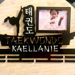 Op de afbeelding: Een zwarte acryl medaillehouder met een silhouet van een persoon die een taekwondo-trap uitvoert, de Koreaanse tekens voor taekwondo, het woord TAEKWONDO, de naam KAELLANIE en een rechthoekige fotolijst met een persoon in een wit uniform.