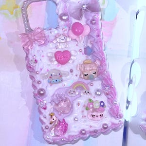 May include: A decorative phone case in shades of pink and white, adorned with various charms. Features include a heart with the word "Love", a swan, a rainbow, and other cute embellishments. The case has a pearl-like texture and a bow.