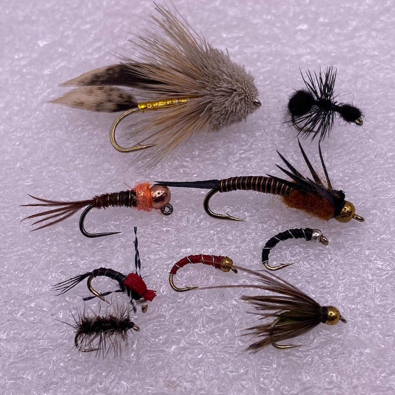 May include: Assortment of eight fly fishing lures with various colors and materials, including brown, black, green, red, and orange.