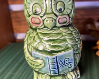 May include: A green ceramic owl figurine holding a book with "ABCs" on the cover. The owl has large eyes, a beak, and small ears. The figurine is on a green surface.