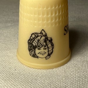 Op de afbeelding: Witte plastic vingerhoed met een zwart-witte illustratie van een meisje met krullend haar. De vingerhoed heeft een getextureerd oppervlak en het woord "Singer" is aan de zijkant gedrukt.