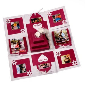 May include: A red and white explosion box with multiple compartments, each containing a photo. The box is decorated with white flowers and a heart-shaped cutout.