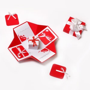 May include: A red and white explosion box with a small white gift box inside. The box has a heart design and is decorated with red and white ribbons. There are also four smaller red squares with white ribbons attached.