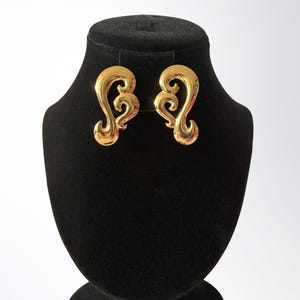 May include: A pair of gold-colored earrings with a swirling, ornate design. The earrings are displayed on a black jewelry stand against a white background. The earrings have a polished, reflective surface.