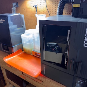 May include: Two 3D printers are on a wooden workbench. The printer on the right is black with the word "ELEGOO" in white. The printer on the left is black with the words "AutoOvertimeDesign ANYCUBIC". An orange tray sits in front.