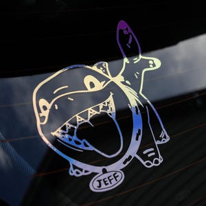May include: A white sticker on a black background featuring a cartoon dinosaur with a large mouth and a tongue sticking out. The dinosaur is wearing a collar with the name "JEFF" written on it.