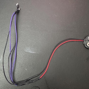 May include: A close-up of a battery holder with two wires attached. The wires are black and red. The battery holder is black and has a silver metal contact point.