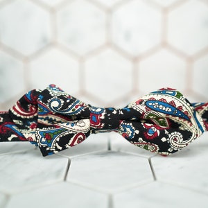 May include: A black paisley bow tie with red, blue, and white accents. The bow tie is made of fabric and is tied in a classic bow shape. The background is a white tile pattern.