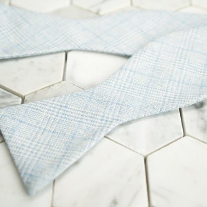 May include: A light blue bow tie with a white and light blue checkered pattern. The bow tie is lying on a white marble surface.