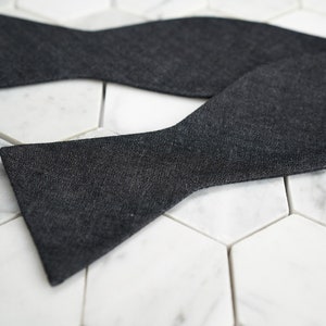 May include: A dark gray bow tie made of a textured fabric. The bow tie is folded and laid on a white marble surface.