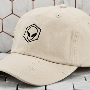 May include: A distressed beige baseball cap with a black alien head embroidered on the front. The alien is inside a hexagon shape.