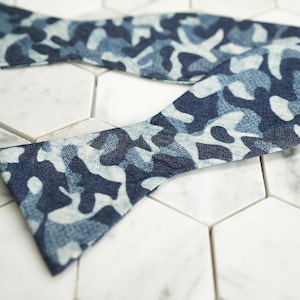 May include: A blue and white camouflage patterned bow tie. The bow tie is made of a soft, textured fabric.