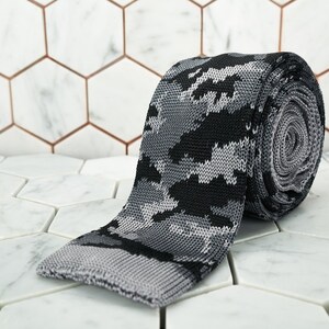 May include: A gray and black knitted tie with a camouflage pattern. The tie is rolled up and lying on a white hexagonal tile surface.