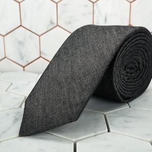 May include: A rolled-up charcoal gray necktie with a subtle textured pattern. The tie is made of a woven fabric and is displayed on a white and gray hexagonal tile surface. The tie is a classic accessory.
