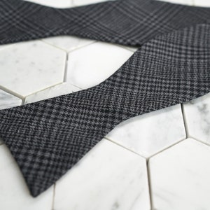 May include: A black and gray bow tie with a houndstooth pattern. The bow tie is made of a soft, textured fabric.