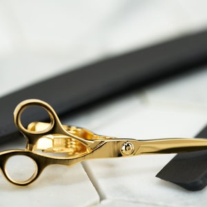 Op de afbeelding: Gouden schaar met een zwart scheermes op een wit marmeren oppervlak.