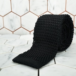 Puede incluir: Una corbata de punto negra enrollada sobre una superficie de baldosas hexagonales blancas.