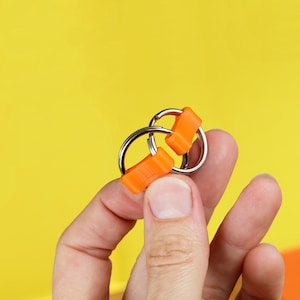 May include: A close-up of a silver key ring with two orange plastic loops. The key ring is being held by a hand.