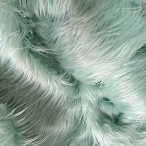 May include: Close-up of a soft, light green faux fur fabric. The fur is long and fluffy, creating a luxurious and plush texture.