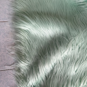 May include: A close-up of a light green faux fur fabric. The fabric has a soft, fluffy texture and is perfect for crafting projects.