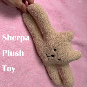 May include: A beige sherpa plush toy in the shape of a bear with black eyes and a black nose. The toy is lying on a pink fuzzy surface. The text "Sherpa Plush Toy" is visible in the image.