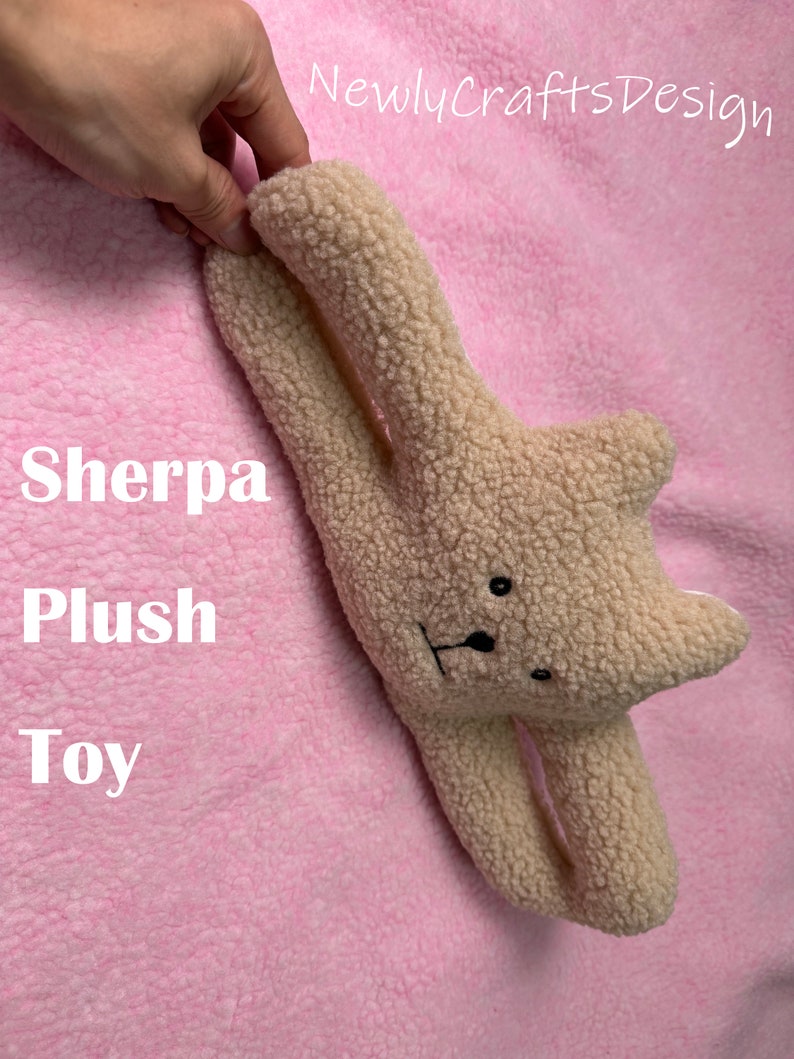 May include: A beige sherpa plush toy in the shape of a bear with black eyes and a black nose. The toy is lying on a pink fuzzy surface. The text "Sherpa Plush Toy" is visible in the image.