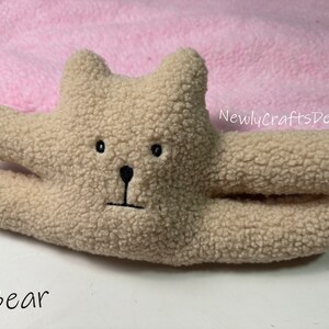 May include: A beige plush toy bear with black embroidered eyes, nose, and mouth. The bear is lying on a pink blanket with the text "NewlyCraftsDesign" in black.