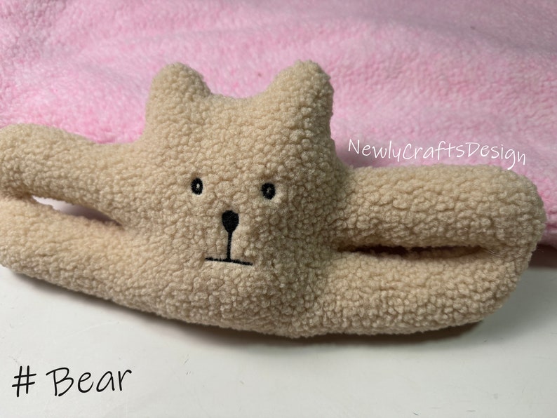 May include: A beige plush toy bear with black embroidered eyes, nose, and mouth. The bear is lying on a pink blanket with the text "NewlyCraftsDesign" in black.