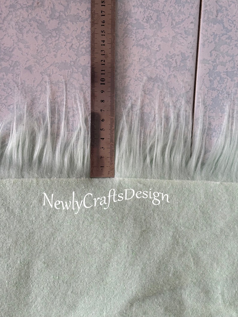 May include: A close-up of a light green fabric with a white faux fur trim. A ruler is visible in the image, showing the fur trim is approximately 7 inches long. The text "Newly Crafts Design" is visible at the bottom of the image.