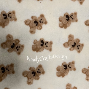 May include: White fabric with a repeating pattern of brown teddy bears. The fabric has a fuzzy texture.