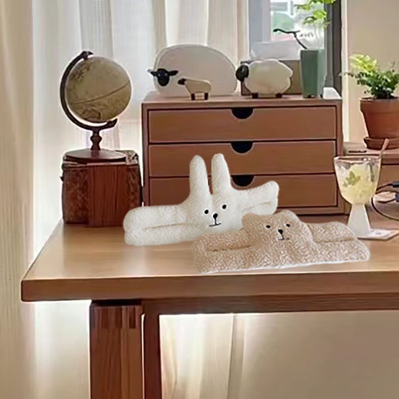 May include: Two plush animal shaped cushions, one white bunny and one beige bear, on a wooden table with a globe, wooden drawers, and a glass of liquid.