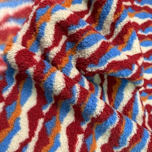 Può includere: Primo piano di un tessuto con motivo rosso, bianco, blu e arancione con una texture soffice. Il tessuto è piegato e mostra il motivo che si ripete.