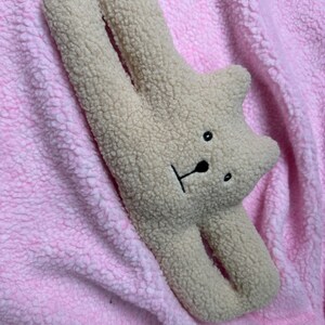 May include: A plush beige bear doorstop with black eyes and a black nose. The bear is lying on a pink fluffy surface. The text "Plush Doors Stopper" is written below the bear.