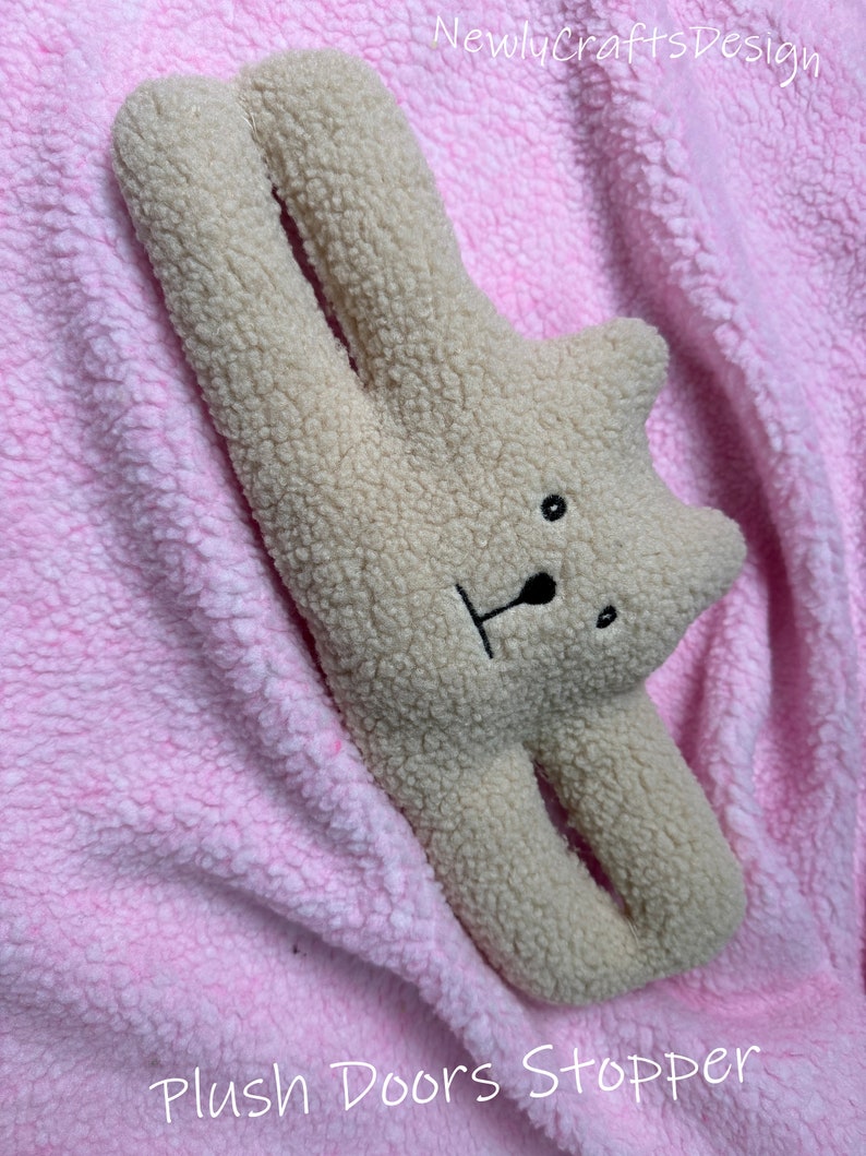 May include: A plush beige bear doorstop with black eyes and a black nose. The bear is lying on a pink fluffy surface. The text "Plush Doors Stopper" is written below the bear.