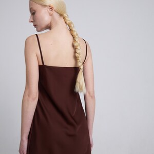 Op de afbeelding: Een vrouw draagt een bruine, mouwloze maxi-jurk met een eenvoudige, vierkante halslijn. Haar blonde haar is gevlochten en in een paardenstaart geknoopt.