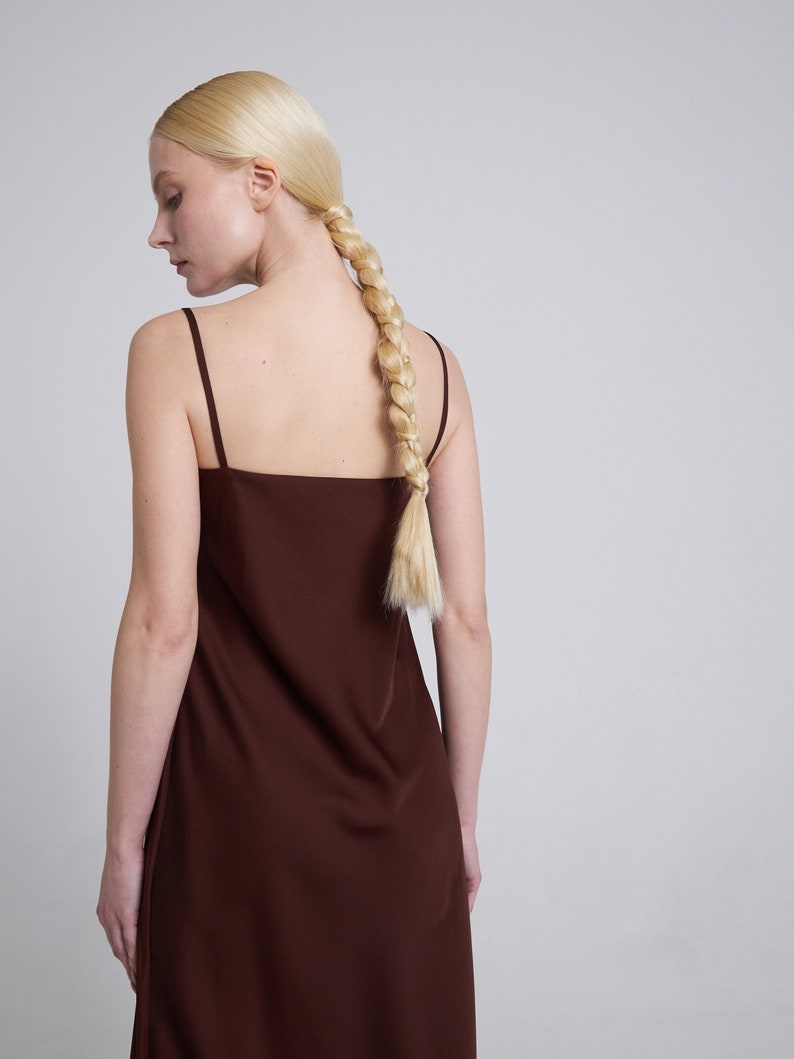 Op de afbeelding: Een vrouw draagt een bruine, mouwloze maxi-jurk met een eenvoudige, vierkante halslijn. Haar blonde haar is gevlochten en in een paardenstaart geknoopt.