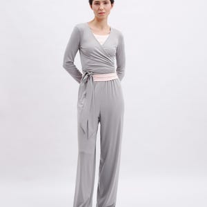 May include: A light gray long-sleeve wrap top with a matching tie waist and wide-leg pants. The outfit features a pale pink undershirt and a pale pink waistband. The ensemble is paired with pale pink ballet flats with small bows on the toes.