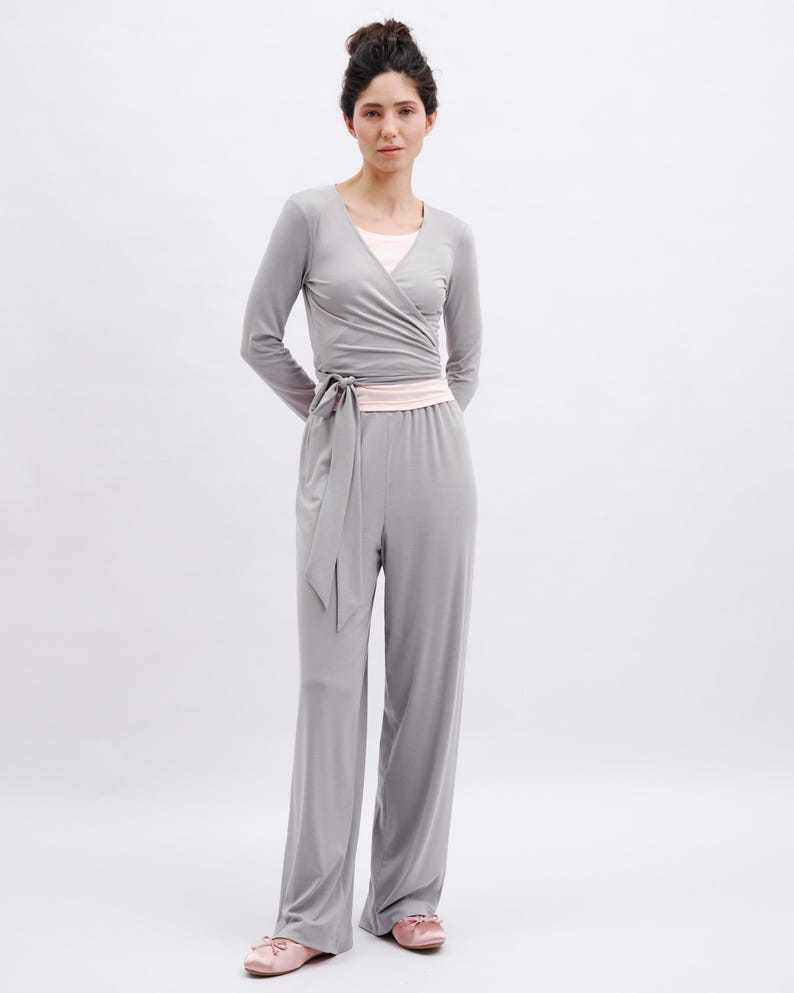 May include: A light gray long-sleeve wrap top with a matching tie waist and wide-leg pants. The outfit features a pale pink undershirt and a pale pink waistband. The ensemble is paired with pale pink ballet flats with small bows on the toes.