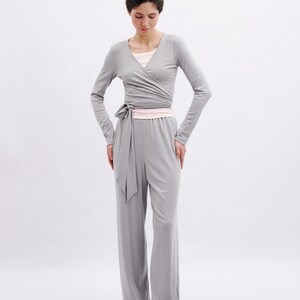 May include: A light gray long-sleeve wrap top with a side tie closure and matching wide-leg trousers. The outfit is layered over a pale pink undershirt and paired with pale pink flat shoes, creating a coordinated ensemble in neutral tones.