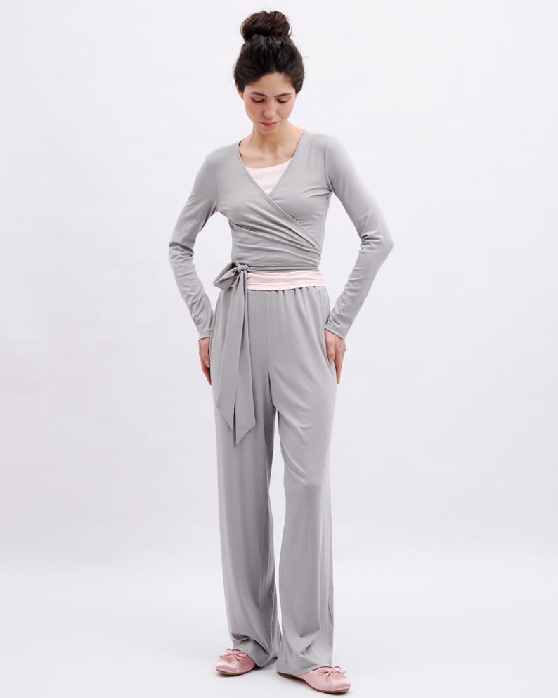 May include: A light gray long-sleeve wrap top with a side tie closure and matching wide-leg trousers. The outfit is layered over a pale pink undershirt and paired with pale pink flat shoes, creating a coordinated ensemble in neutral tones.