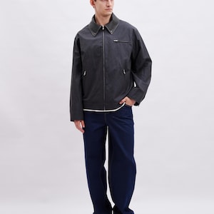May include: A dark gray jacket with a zipper closure and a dark green collar. The jacket is paired with dark blue jeans and black sneakers. The person is standing in front of a white background.