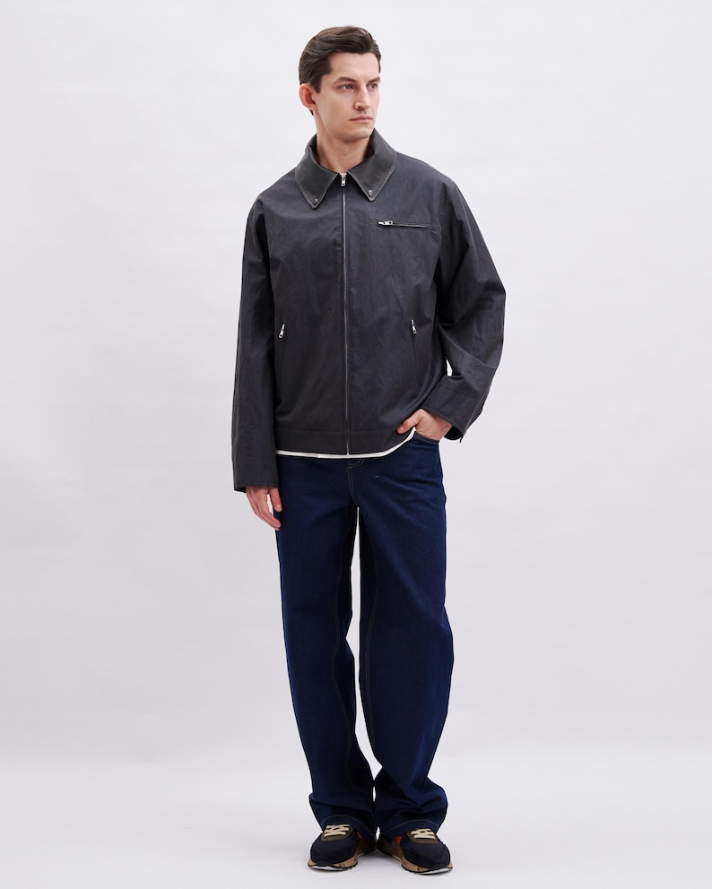 May include: A dark gray jacket with a zipper closure and a dark green collar. The jacket is paired with dark blue jeans and black sneakers. The person is standing in front of a white background.