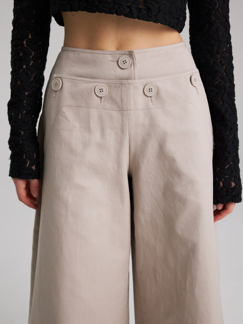 May include: Beige wide-leg cropped pants with a high waist and multiple button details.