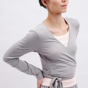 May include: A long-sleeved gray wrap top featuring a V-neckline and a side tie closure. The garment is layered over a light pink scoop-neck undershirt, creating a dual-tone appearance with fitted sleeves and a soft, smooth fabric texture throughout.